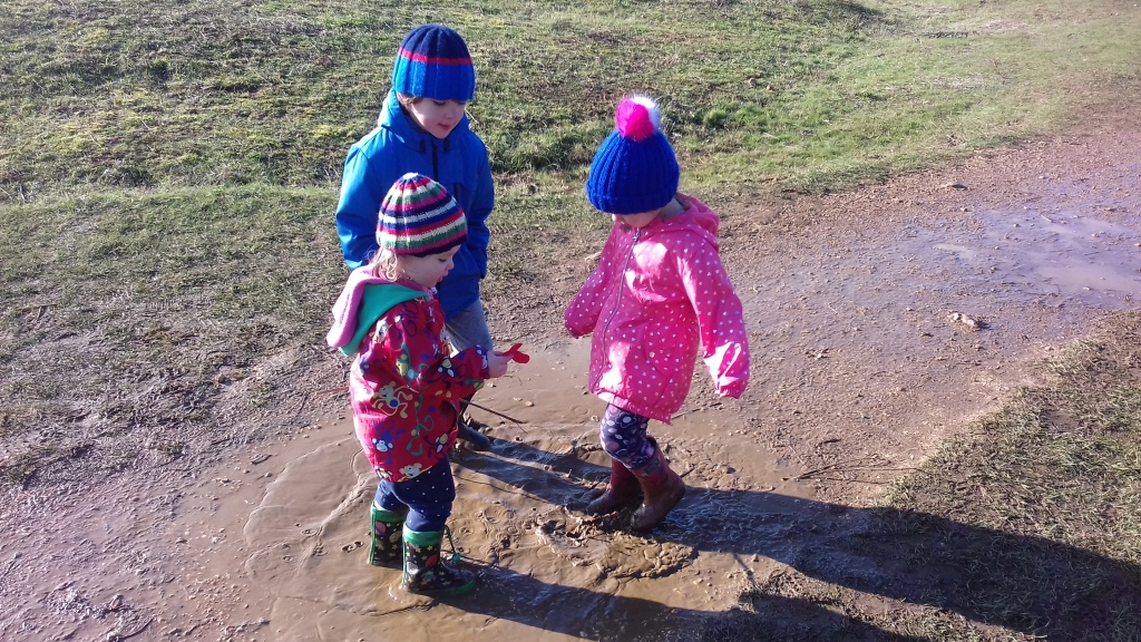 Three kids with hats and coats around a muddy puddle